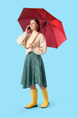 Sonbahar elbiseli, şemsiyeli, mavi arka planda cep telefonuyla konuşan kadın turist.