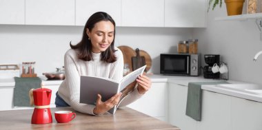 Bir fincan espresso ve gayzer kahve makinesiyle genç ve güzel bir kadın modern mutfakta dergi okuyor.