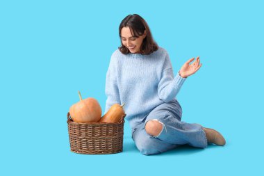 Mavi arka planda oturan sepet dolusu balkabağı olan genç kadın.
