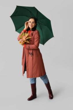 Gri arka planda sonbahar yaprakları ve şemsiyesi olan güzel genç bir kadın.