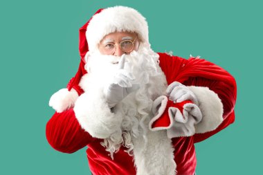 Bir çanta dolusu Noel hediyesiyle Noel Baba turkuaz arka planda sessiz bir jest sergiliyor.