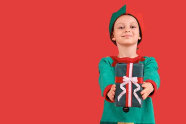 Kırmızı arka planda Noel hediyesi olan elf kostümlü şirin bir çocuk.