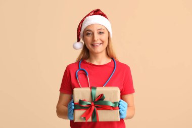 Noel Baba şapkalı olgun kadın doktor. Bej arka planda Noel hediyesi kutusu var.