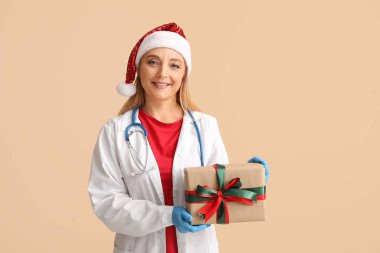 Noel Baba şapkalı olgun kadın doktor. Bej arka planda Noel hediyesi kutusu var.