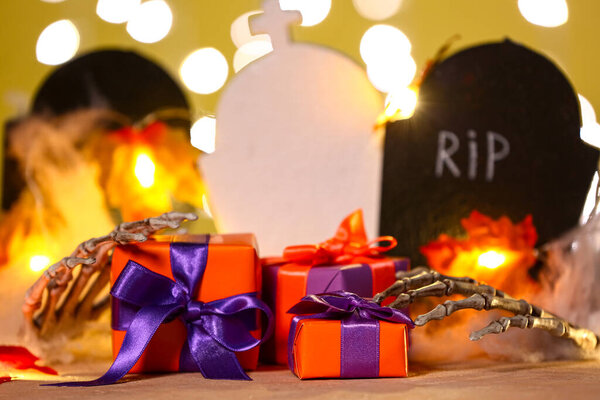 Composition with beautiful gift boxes, skeleton hands and graves for Halloween on table against blurred lights, closeup