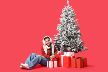 Kırmızı arka planda Noel ağacının yanında hediye kutuları olan güzel genç bir kadın.