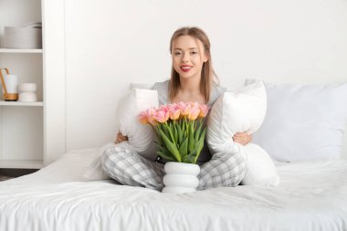 Yatak odasında bir buket lale çiçeğiyle rahat bir yatakta oturan mutlu genç kadın.