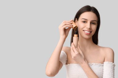 Beautiful young woman opening foundation on grey background