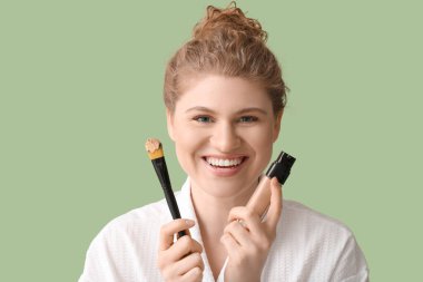 Yeşil arka planda fondöten şişesi ve makyaj fırçası olan mutlu genç bir kadın.