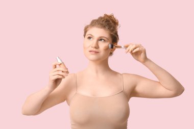 Pembe arka planda fondöten şişesi ve makyaj fırçası olan güzel bir genç kadın.