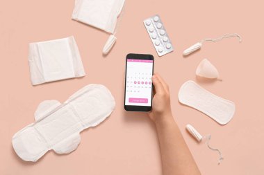 Cep telefonu ekranında regl takvimi olan kadın eli ve bej arka planda kadın hijyen ürünleri.