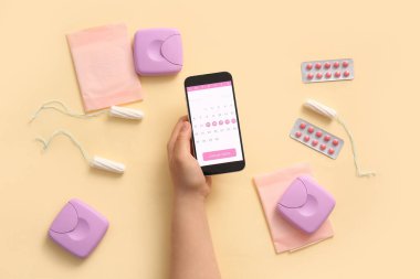 Cep telefonu ekranında regl takvimi olan kadın eli ve bej arka planda kadın hijyen ürünleri.