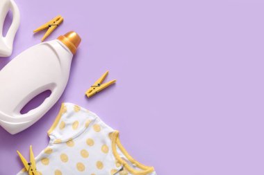 Çamaşır deterjanı, mandallar ve mor arka planda bebek kıyafeti.