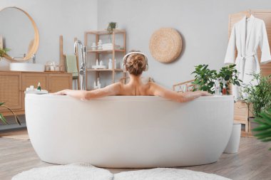 Kulaklıklı genç bir kadın evde banyo yapıyor, arkadan bakıyor.