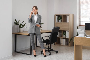 Güzel genç iş kadını cep telefonuyla konuşuyor ve modern tablet bilgisayarını ofisinde tutuyor.