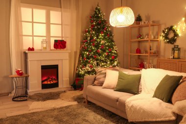 Şenlikli oturma odasının içinde şömine, Noel ağacı ve akşamları parlayan ışıklar.