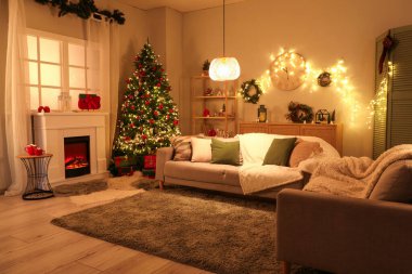 Şenlikli oturma odasının içinde şömine, Noel ağacı ve akşamları parlayan ışıklar.