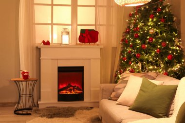 Şenlikli oturma odasının içinde şömine, Noel ağacı ve akşamları parlayan ışıklar.