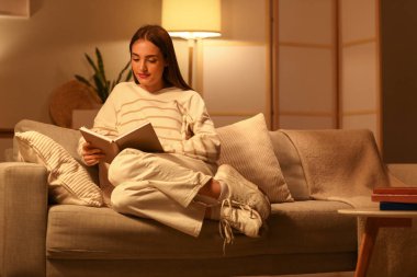 Evdeki kanepede kitap okuyan güzel genç bir kadın.