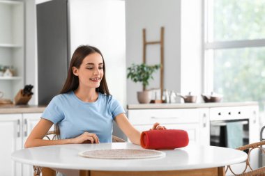 Güzel, genç, mutlu, kablosuz hoparlörlü bir kadın mutfaktaki masada oturuyor.