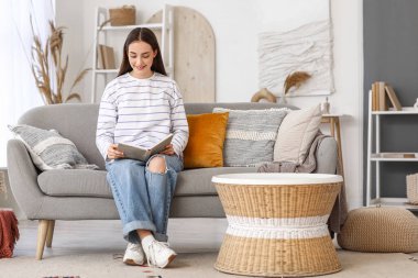 Evdeki kanepede kitap okuyan genç ve güzel bir kadın.