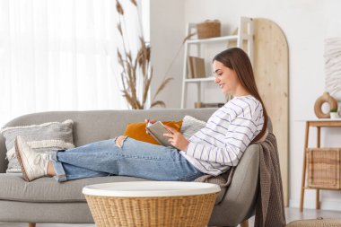 Evdeki kanepede kitap okuyan genç ve güzel bir kadın.