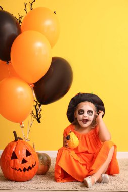 Cadılar Bayramı için giyinmiş, sarı duvarın yanında balon ve balonlarla şaşırmış küçük bir kız.
