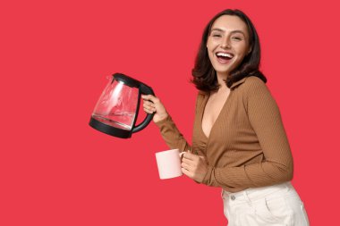 Kırmızı arka planda modern elektrikli su ısıtıcısı ve fincanı olan güzel genç bir kadın.