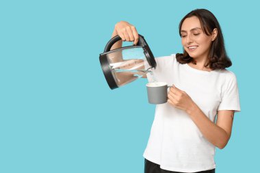 Güzel genç bir kadın modern elektrikli çaydanlıktan sıcak suyu mavi arka planda kupaya döküyor.