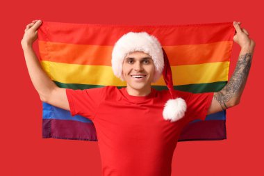 Kırmızı arka planda LGBT bayrağı olan Noel Baba şapkalı genç adam.