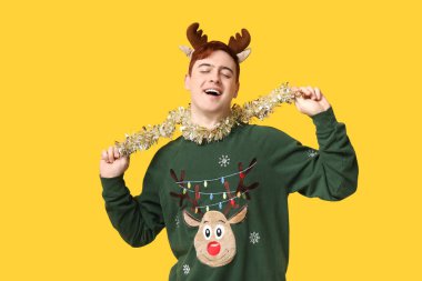 Sarı arka planda kalın süveter, süs ve ren geyiği saç bandı takan yakışıklı bir genç.