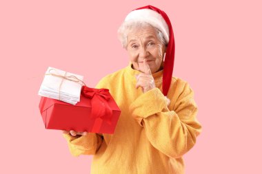 Noel Baba şapkalı yaşlı bir kadın. Elinde mektuplar ve Noel hediyesi var. Pembe arka planda sessizlik jesti yapıyor.