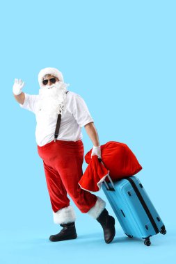 Noel Baba bir çanta dolusu hediyeyle ve mavi arka planda el sallayan bavulla