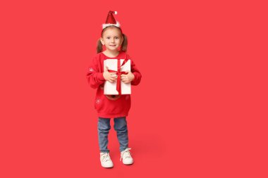 Kırmızı arka planda hediyeli Noel süveteri içinde şirin küçük bir kız.