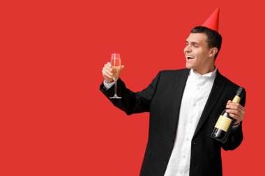 Parti şapkalı genç adam kırmızı arka planda bir şişe şampanya ile Noel 'i kutluyor.