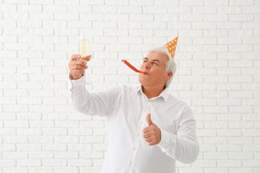 Parti şapkalı son sınıf öğrencisi bir bardak şampanyayla Noel 'i kutluyor ve beyaz tuğla arka planda baş parmak hareketi yapıyor.