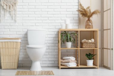 İçinde seramik klozet, katlanmış ekran, örülmüş sepet ve beyaz tuğla duvarın yanında raf ünitesi olan şık bir tuvalet.