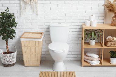 Seramik klozetli şık tuvaletler, örülmüş sepet ve beyaz tuğla duvarın yanındaki raf ünitesi.