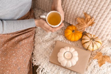 Elinde bir fincan yeşil çay olan kadın oturma odasında kitap, mum ve balkabağı yanında oturuyor. Yakın çekim.