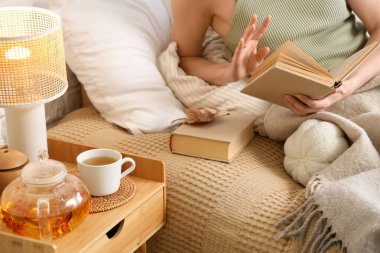 Çaydanlık ve çay yatak odasında kitap okuyan bir kadının yanındaki komodinin üzerinde. Yakın çekim.