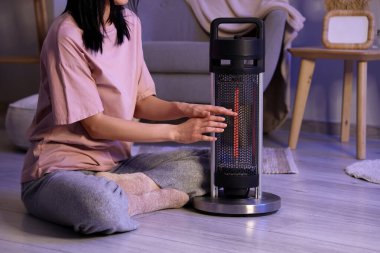 Genç bir kadın akşamları elektrikli ısıtıcının yanında ellerini ısıtıyor.