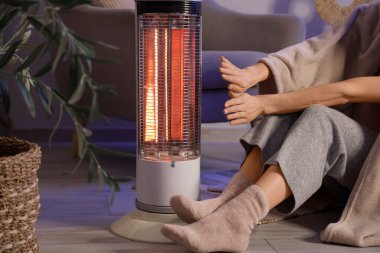 Elleri elektrikli ısıtıcının yanında ısıtan battaniyeli genç bir kadın akşam evde, yakın çekim yapıyor.