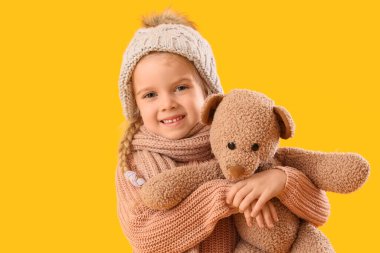 Sarı arka planda oyuncak ayısı olan, kışlık giysili şirin küçük kız.