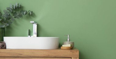 Banyoda yeşil duvarın yanında beyaz lavabo ve banyo malzemeleri var. Tasarım için pankart
