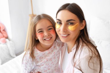 Happy mother and her little daughter with under-eye patches in bedroom, closeup