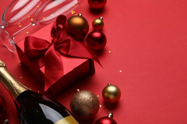 Bir şişe şampanya, hediye kutusu ve kırmızı arka planda Noel süslemeleri.
