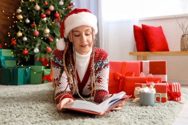 Noel Baba şapkalı genç bir kadın Noel arifesinde evde kitap okuyor.