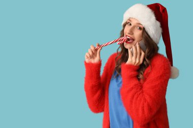 Noel Baba şapkalı genç kadın cep telefonuyla konuşuyor ve mavi arka planda şeker kamışı yiyor.