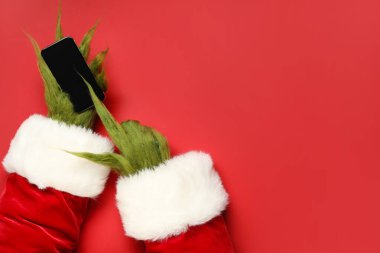 Green hairy hands of creature in Santa costume with mobile phone on red background clipart