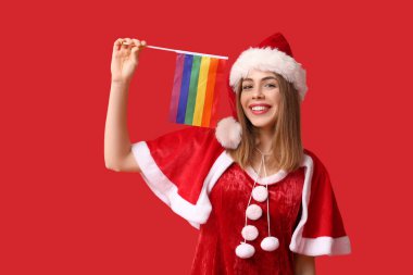 Kırmızı arka planda LGBT bayrağı olan Noel Baba kostümlü genç bir kadın.